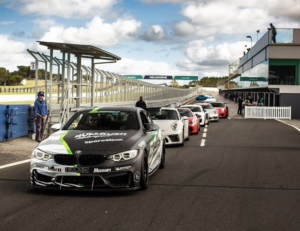 A BMW M4 race car leads a lineup of performance cars with custom car wraps at a racetrack, featuring a custom Bullrush Rally wrap.